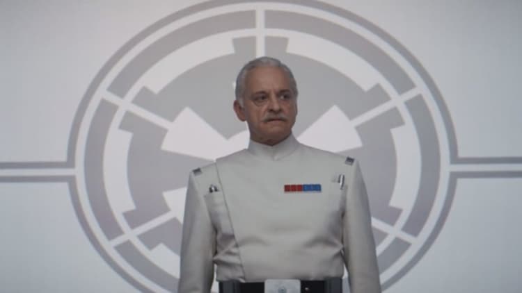 Colonel Wullf Yularen stands in front of a large Imperial logo. He is dressed in a white Imperial Officer uniform.