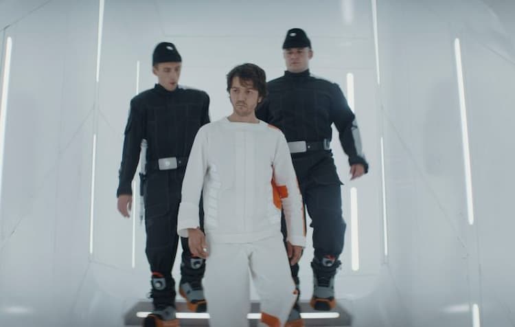 Cassian, dressed in prison clothes, is escorted by two Imperial prison guards down a pristine white corridor.