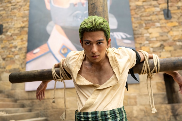 Zoro is tied up on a cross in a courtyard. A large picture of Morgan can be seen on the wall behind him.