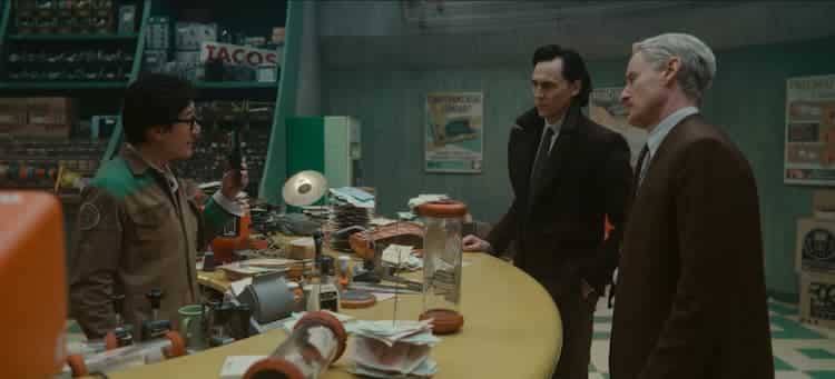 Loki and Mobius are talking with O. B. at his desk. The desk is covered in various items and tubes. An assortment of posters are in the background.
