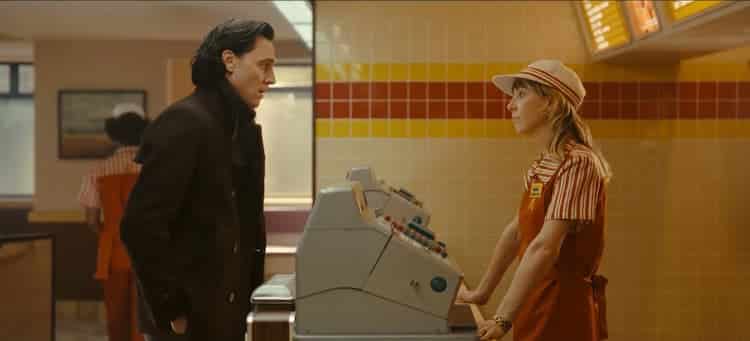 Loki and Sylvie are in a McDonald’s. Loki is wearing a dark coat and Sylvie is wearing an employee uniform. A line of registers separate them.