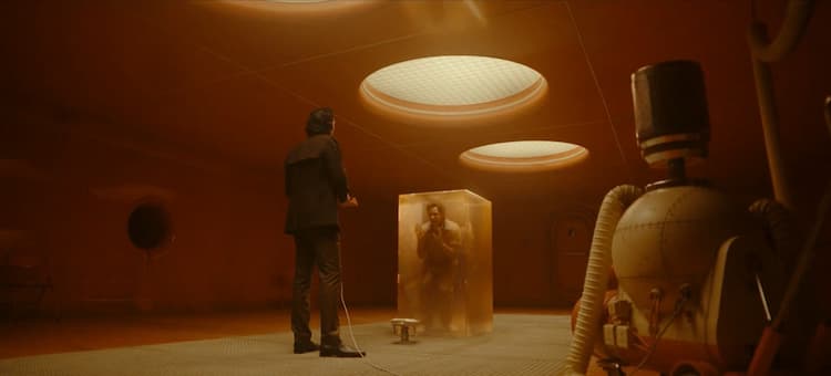 Loki is standing in front of Brad who is trapped in a translucent block.There's a machine to the right and some light from above. The room is an orange colour.
