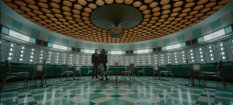 Loki and Sylvie are standing in a room with walls lined with pie cabinets. The room is mostly green with yellow lights above. Tables and chairs are peppered throughout the room