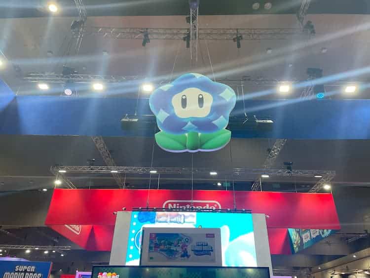 A Wonder Flower is hanging from the ceiling. A screen playing a Mario trailer is below it. The red Nintendo banner can also be seen behind the flower.