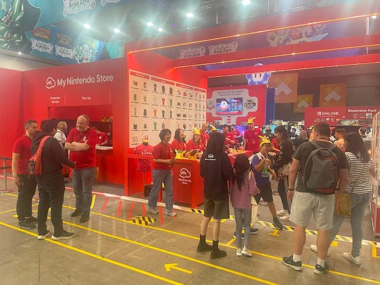 The My Nintendo Store. The store is a bright red colour with a large image on a wall that showcases all the products available to purchase. Nintendo staff in red shirts are scattered around the area. People are lining up to enter the store as well.