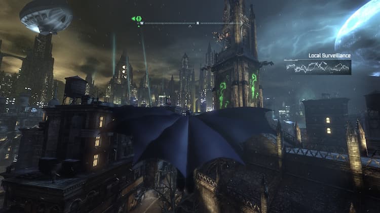 Batman is gliding through Arkham City. There are Riddler markings on the church to the right. The compass is at the top of the screen. Local surveillance is being picked up on the top right of the screen.