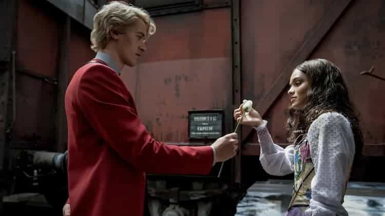 Snow is handing Lucy Gray a white rose. Snow is wearing a red uniform and Lucy Gray is wearing a brightly coloured dress. They are standing in front of a reddish train car.