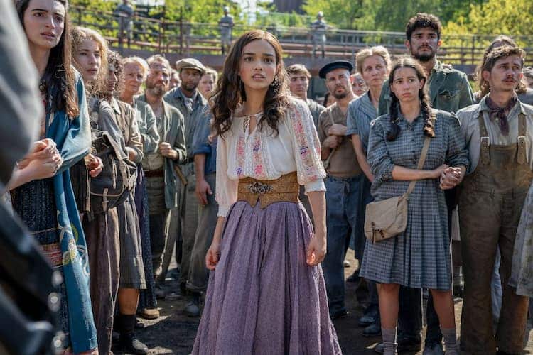 Lucy Gray is standing in the middle of a group of other civilians. Everyone has worried looks on their faces. Guards can be seen in the background above everyone else.