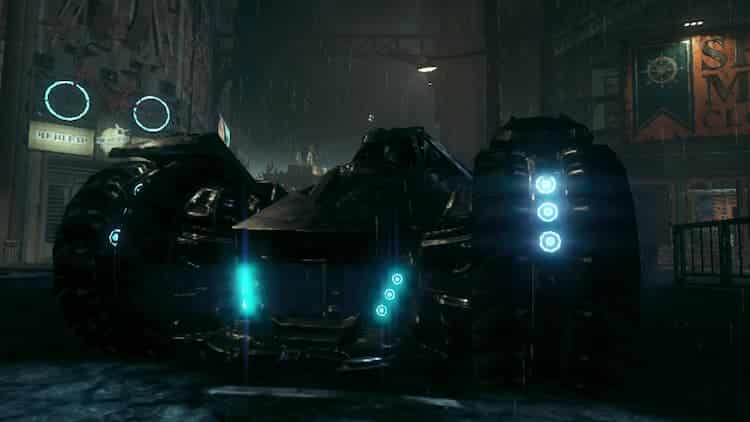 Batman is inside the Batmobile. The headlights of the Batmobile are blue. It's raining and various storefronts are behind the Batmobile.