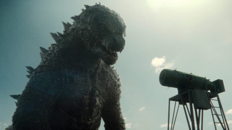 Godzilla is standing and staring at an atomic bomb. The bomb is on a metal tower. The sky is clear behind Godzilla.