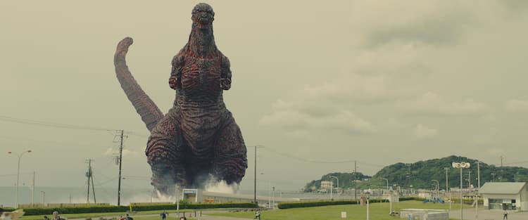 Godzilla is standing in the water just in front of the grass. Power lines and buildings are on the right. Some mountains are in the background.