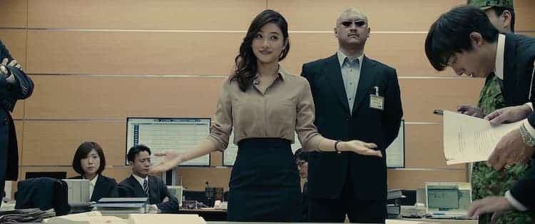 Kayoko has her arms in an open position. Her bodyguards is standing behind her. Various people are standing or sitting around the room. Computer screens surround the room.