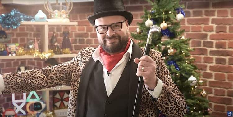 Robert S. Pearson wears a tophat, a leopard print jacket and a red cravat. He is holding a cane in his left hand. He’s standing in front of a christmas tree and PlayStation themed shelving