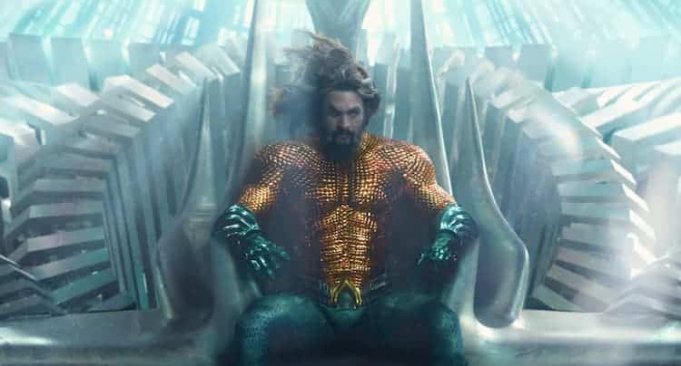 Aquaman is sitting on the throne of Atlantis. The throne is white and the surrounding room is also white. Light is coming in from the top of the screen.