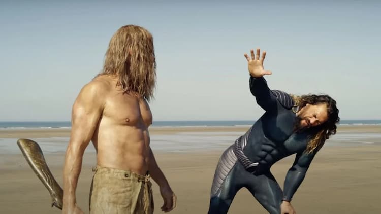 Aquaman is raising his hand towards Orm. Aquaman is wearing a stealth suit and is hunched over. Orm has long hair and a beard and is also shirtless. The two are standing on a beach.