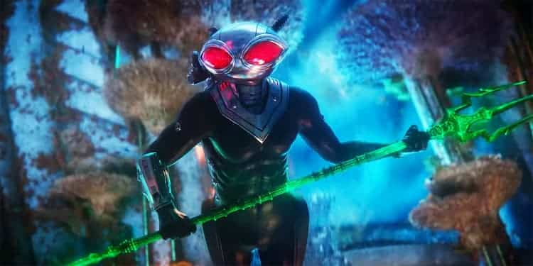 Black Manta is in the ocean while holding a black trident that is glowing green. Various marine life is behind him.