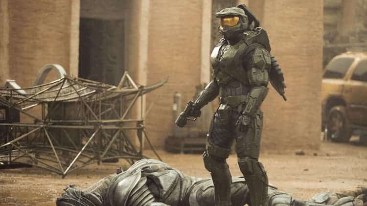 Master Chief is standing over an Elite. Chief has his gun pointed at the Elite. The Elite is dead and the Chief is looking in a different direction. They are in a town covered in sand.