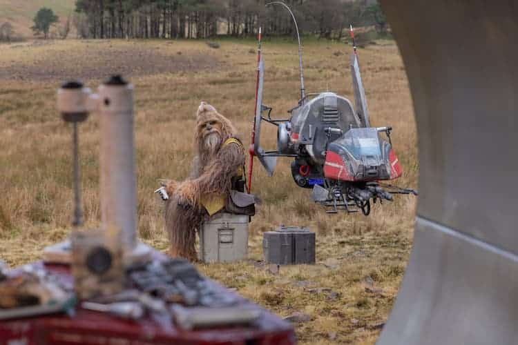 Kelnacca is sitting on a box next to a speeder bike in a field. Various pieces of equipment are scattered around the area. The tree line is in the background. Kelnacca is looking towards the camera.