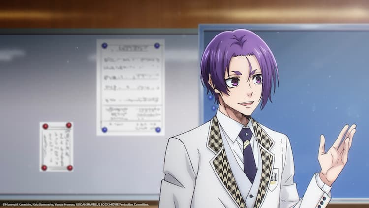 Reo is standing in a classroom. He has his left hand raised and is talking to someone. A whiteboard with two pieces of paper stuck on it is in the background as well as a blank chalkboard.